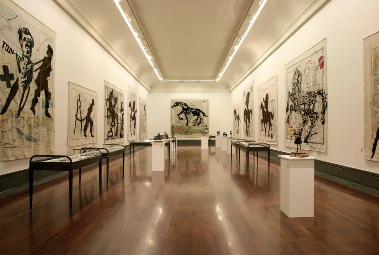 Heller Ausstellungsraum mit Wandteppichen, Skulpturen auf Podesten und Vitrinen, glänzender Holzfußboden